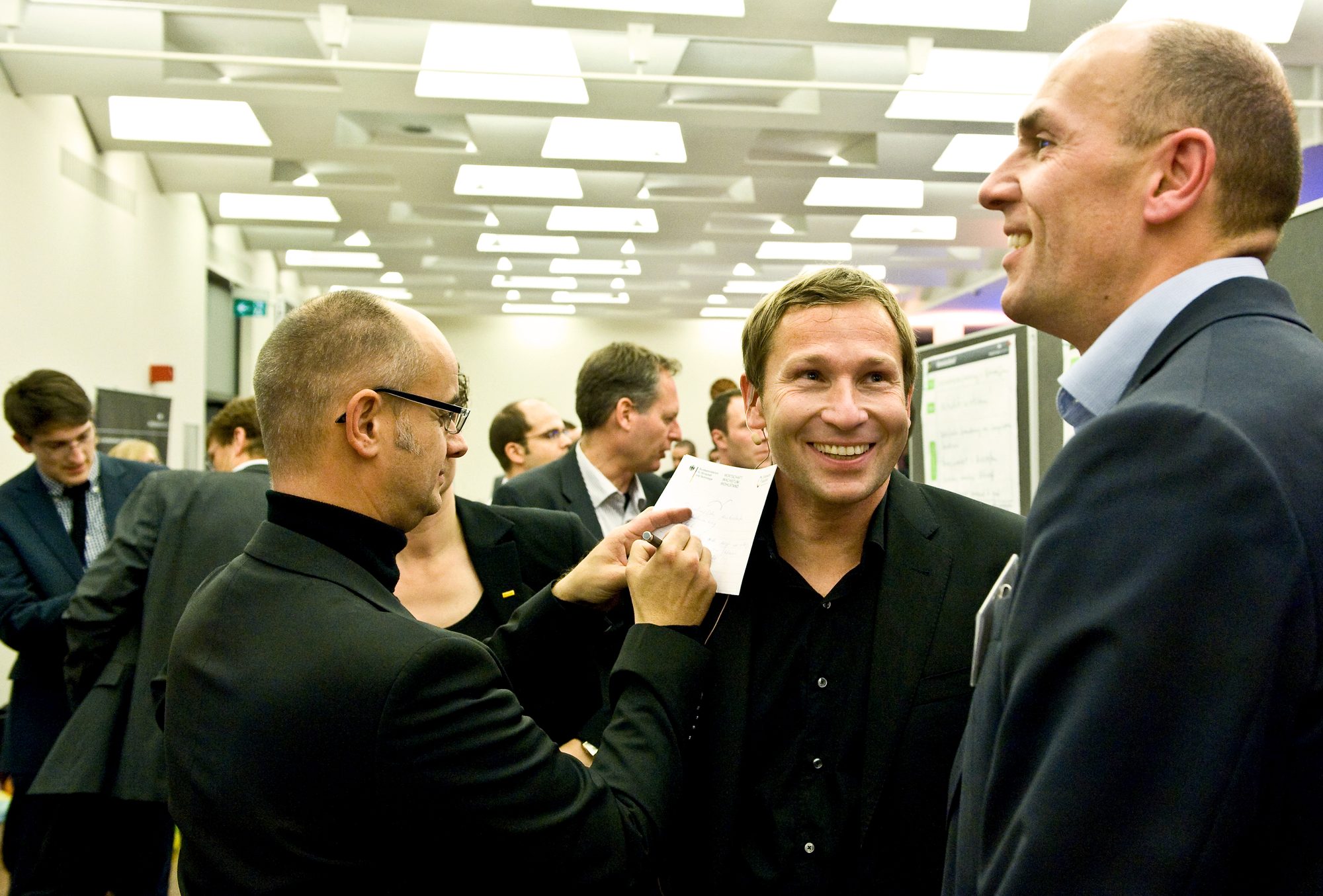 Three participants in conversation at a business event, one of them noting something down